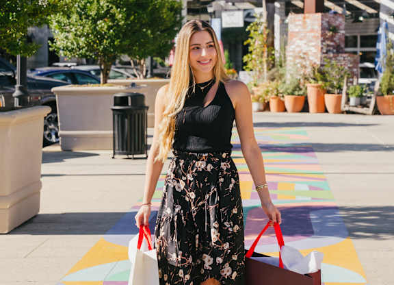 A woman in a black top and floral skirt is walking with shopping bags.