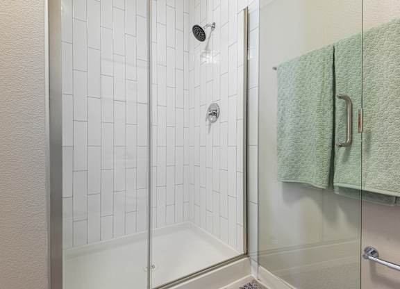 A white tiled shower with a glass door and a green towel hanging on the wall.