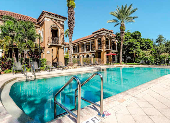 Sparkling swimming pool framed by palm trees and Mediterranean-style buildings.