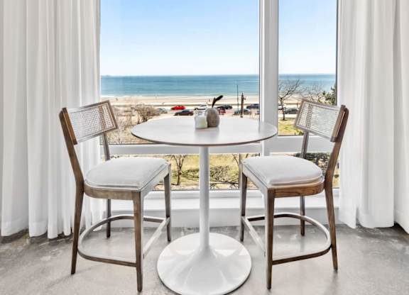 A dining table with chairs is set up by a window with a view of the beach.