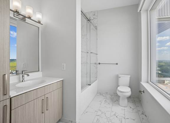 Penthouse bathroom with a glass-enclosed tub and shower, a quartz vanity, marble-look tile flooring and a window.