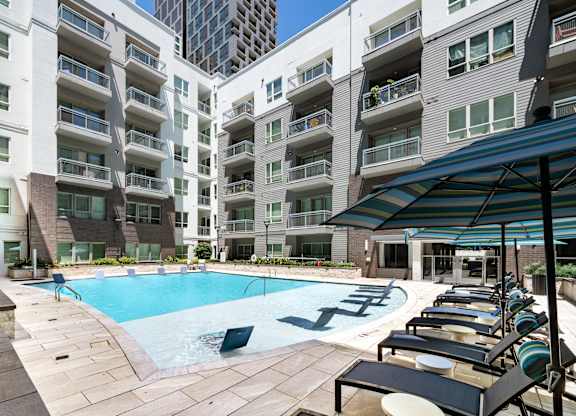 A swimming pool surrounded by lounge chairs and umbrellas in front of apartment buildings.