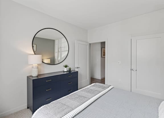 Apartment bedroom with a bed, dark dresser, round mirror, and two interior doors.