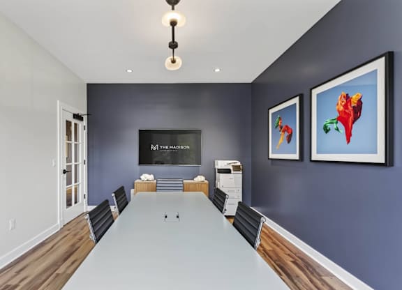 Meeting room with a long white table, black chairs, artwork, and a wall screen.