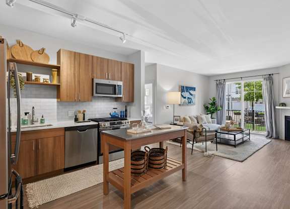 Open apartment kitchen and living room with stainless steel appliances, island, sofa, and patio door.