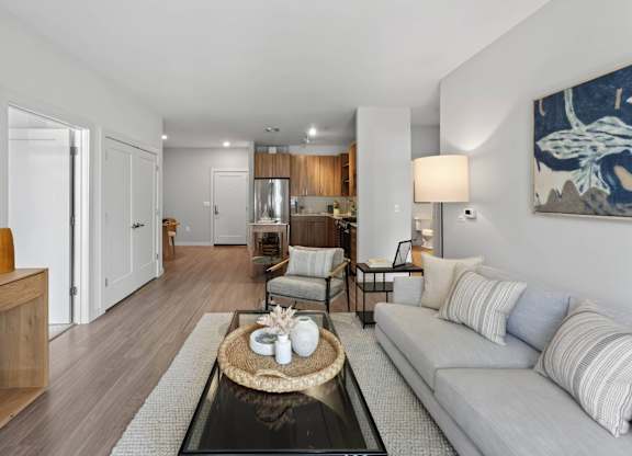 Open apartment living area with a sofa, coffee table, kitchen, and wood-style flooring.