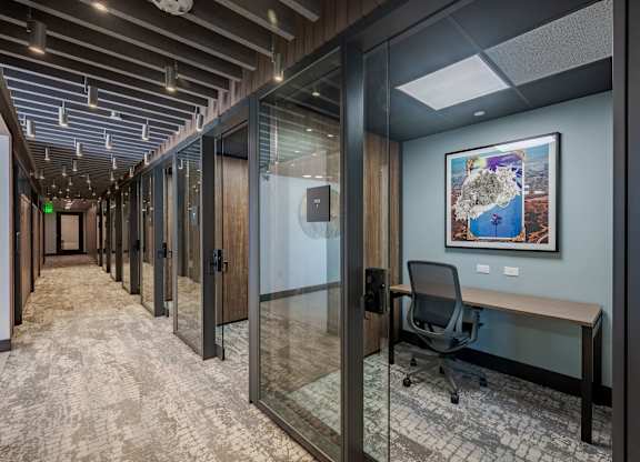 Coworking corridor with glass-enclosed offices, desks, wood doors, and overhead lighting.