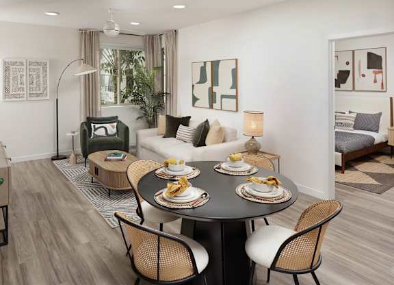 Dining area beside a furnished living room with a bedroom doorway and large windows.