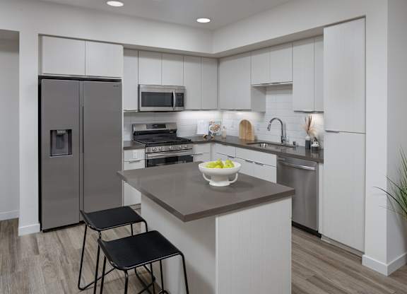 Kitchen with gray cabinetry, quartz island, stainless steel appliances, and two counter stools.