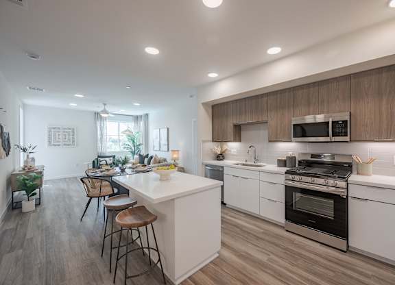 Open kitchen with a quartz island, counter stools, stainless steel appliances, and living space beyond.