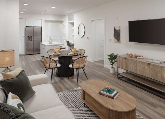 Living room with a sofa, coffee table, dining area, wall-mounted television, and kitchen beyond.