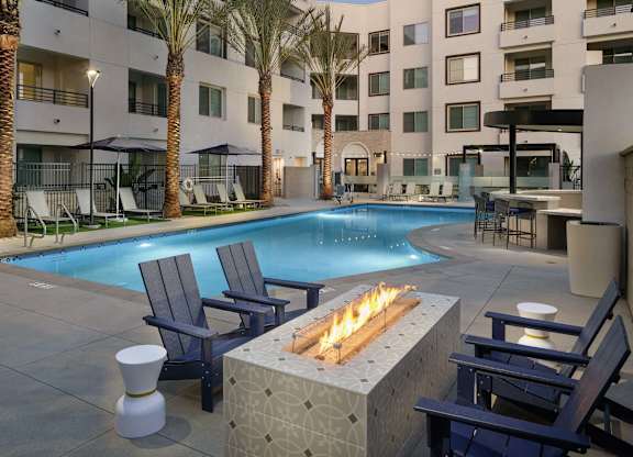 Pool courtyard at dusk with lounge chairs, palm trees, apartment balconies, and a linear fire pit.
