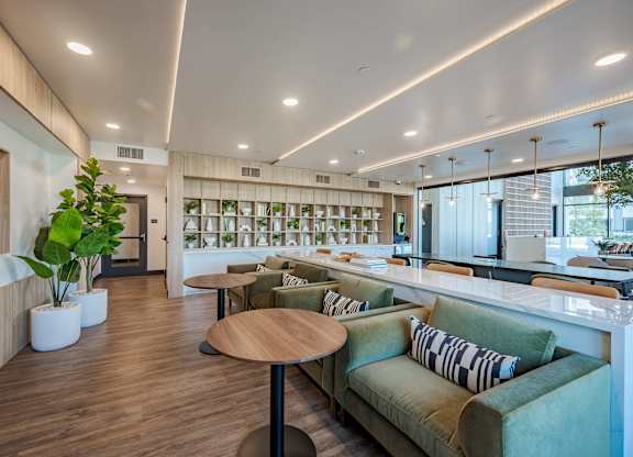 Resident lounge with sectional seating, round tables, bookshelves, plants, and floor-to-ceiling windows.