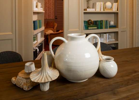 A white jug sits on a table in front of a shelf with books and other items.