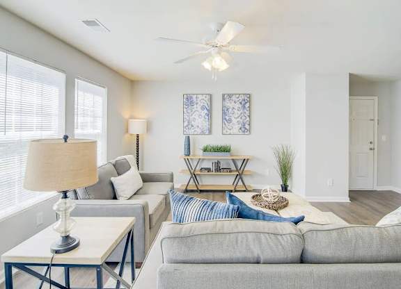 A living room with a grey couch, a table with a lamp, and a ceiling fan.