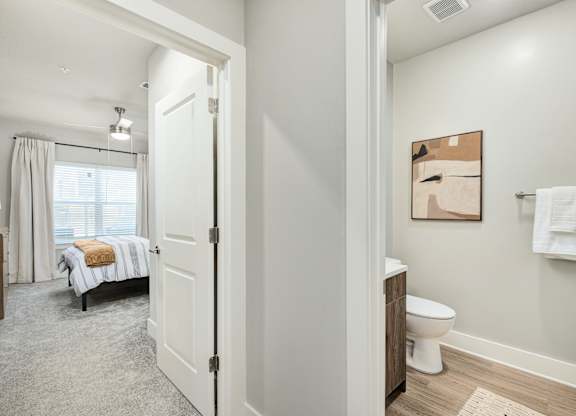 A white bathroom with a toilet, sink, and a bedroom with a bed and window at The Ashton at East Park Apartments, Kennesaw, Georgia