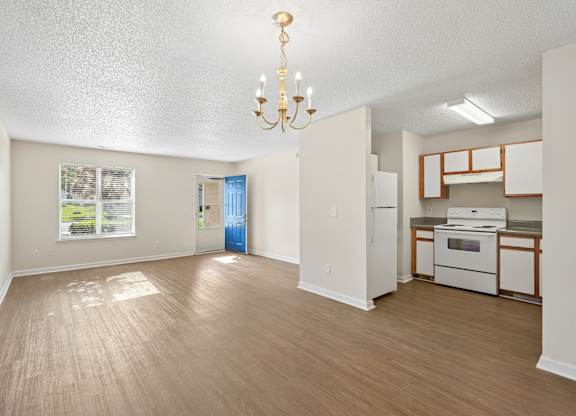 A well-lit, empty room with a chandelier and a kitchenette in the background.