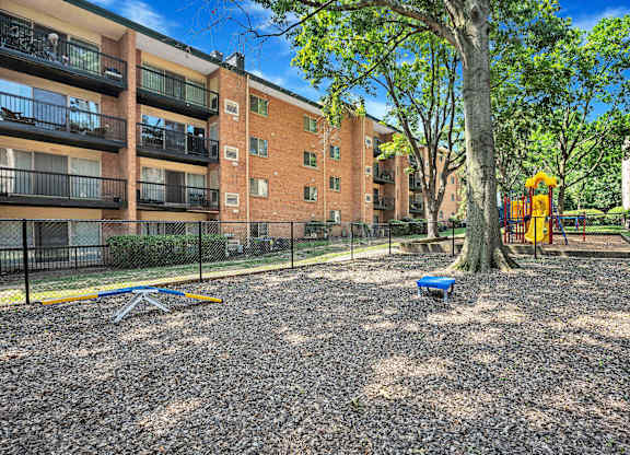 A playground area with a slide, swings, and a tree in front of a brick apartment building.
