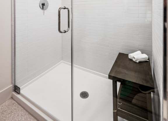 A white tiled shower with a glass door and a black stool.