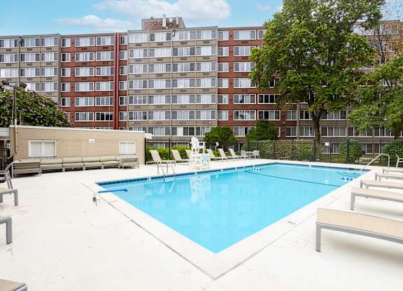 A large swimming pool surrounded by lounge chairs and trees in front of apartment buildings.