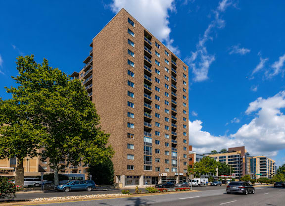 A tall brown building with many windows is in the middle of a street with cars and trees.