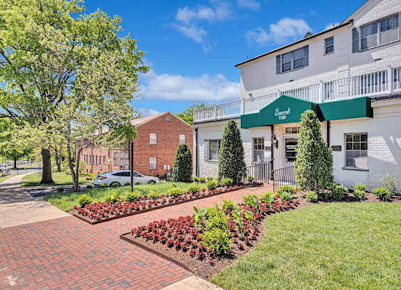 A white building with a green awning is surrounded by a brick walkway and a garden with red flowers.