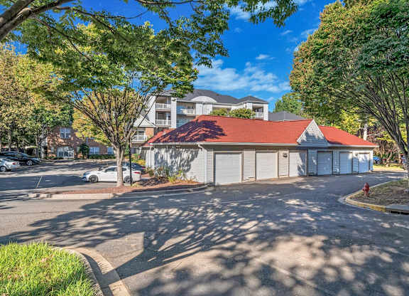 A white house with a red roof is surrounded by trees and a parking lot.