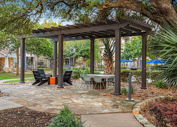 A gazebo with a table and chairs is surrounded by plants and trees.