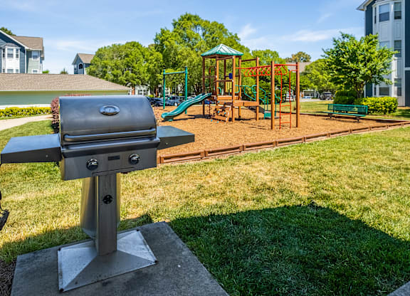 A barbecue grill is in the foreground of a park with a playground in the background.