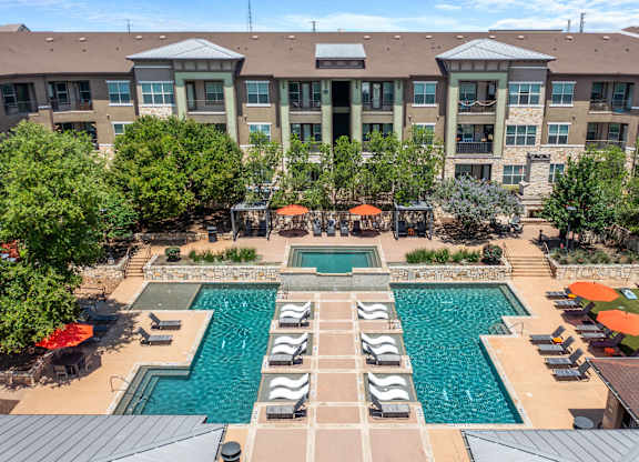 A large swimming pool surrounded by lounge chairs and umbrellas in front of a multi-story apartment building.