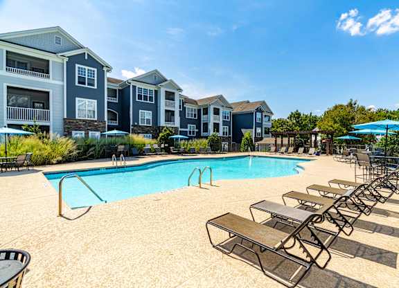 A pool surrounded by lounge chairs and umbrellas in front of apartment buildings.