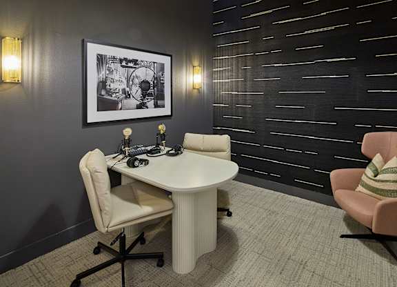 A black and white picture hangs on a dark wall above a white table with a laptop and a chair with a striped pillow on it.