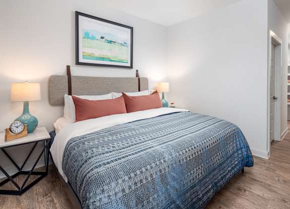 A bed with a blue blanket and red pillows is in the foreground of a room with a painting on the wall and a nightstand with a clock and a vase.