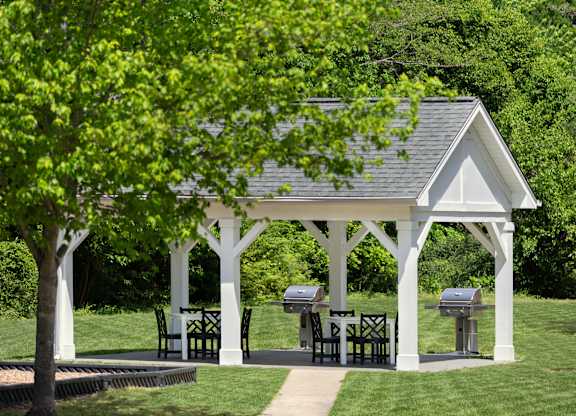 A white gazebo with a table and chairs is surrounded by greenery.