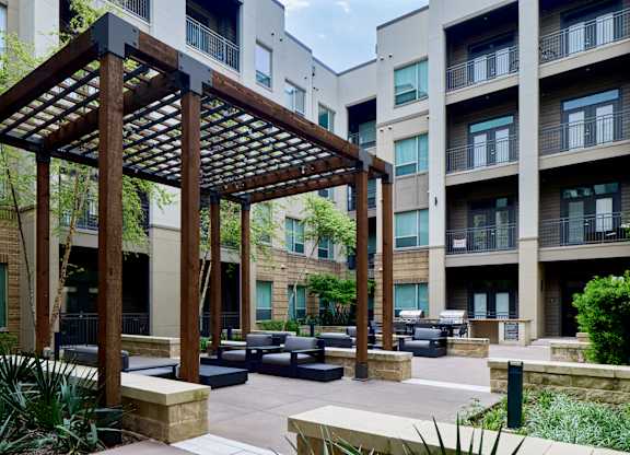 A wooden pergola is in the middle of a courtyard with black seating.