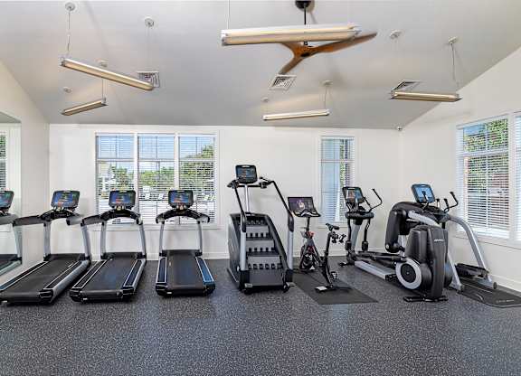 A row of treadmills and exercise bikes are lined up in a brightly lit gym.