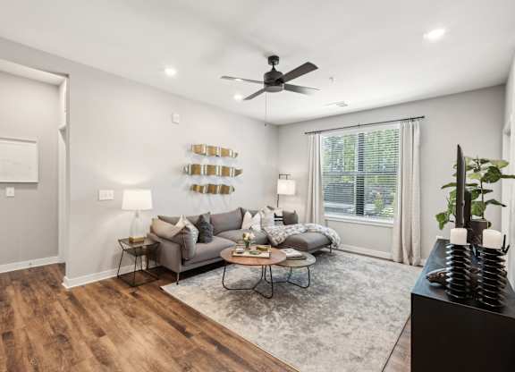 A living room with a grey couch, a coffee table, a ceiling fan, and a large window.