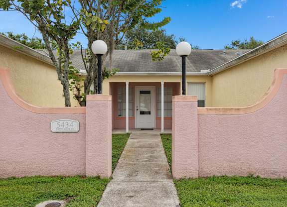 the front entrance to a pink house with a sidewalk and street lamps at The Groves at Wimauma Apartments , Florida 33598