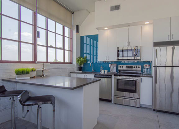 Steelcote Lofts kitchen with Steelcote blue backsplash, gray quartz countertops, stainless steel appliances, white cabinetry, kitchen island and polished concrete floors at Steelcote Square, St. Louis