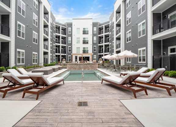 A pool surrounded by lounge chairs and umbrellas in front of apartment buildings at The Oliver Apartments, Olivette, MO