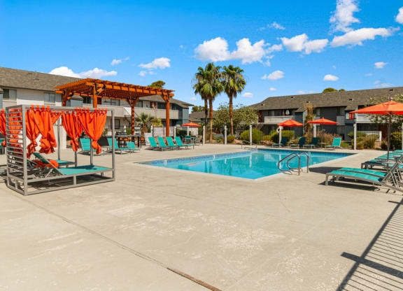 Apartments in West Las Vegas, NV - Accent on Rainbow - Resort Style Swimming Pool With Teal Lounge Chairs, Orange Umbrellas, And Palm Trees Under Blue Sky