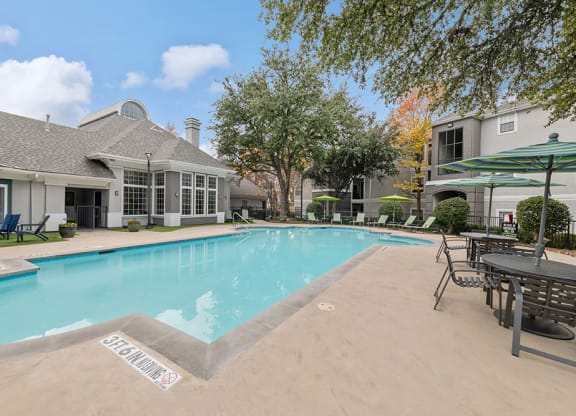 2-Bedroom Apartments in Lewisville, TX - Bridges at Oakbend - Outdoor Pool With Lounge Chairs, Patio Tables With Umbrellas, And Clubhouse Surrounded By Trees.