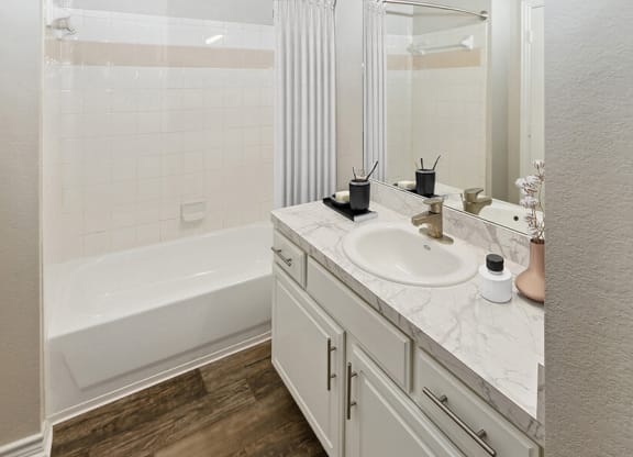 Model Bathroom with White Cabinets and Wood-Style Flooring at Belmont at Duck Creek Apartments in Garland, TX.