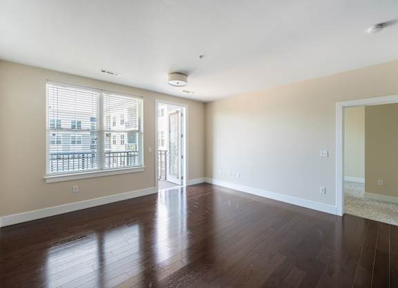 A large empty room with wooden floors and a view of a balcony through the window.