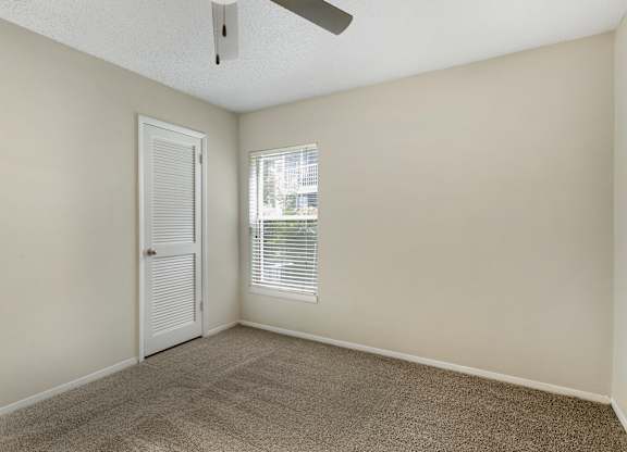 A room with a carpeted floor, a ceiling fan, and a window with blinds.