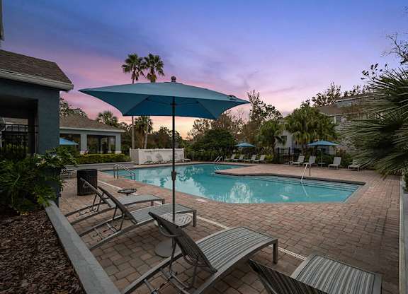 Westside Jacksonville, FL, Apartments Near Oakleaf Town Center - Westland Park - A Swimming Pool Area Features Lounge Chairs, Blue Umbrellas, And Palm Trees.