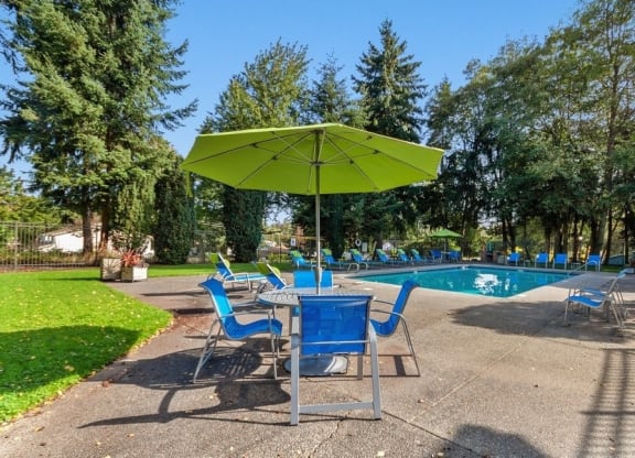 Federal Way, WA, Apartments Near Costco - Sunset Three Twenty - A Swimming Pool With Blue Lounge Chairs, Concrete Deck, Green Umbrella Seating Area, And Tall Evergreen Trees.