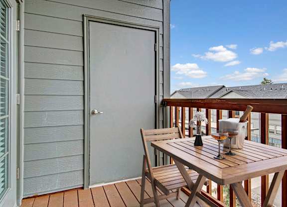 A wooden table with two chairs is on a deck with a white door on a patio.