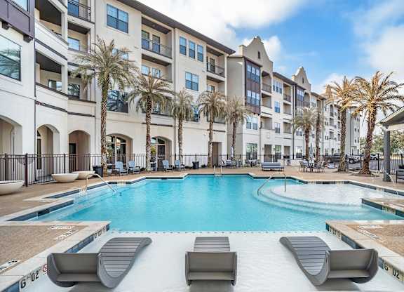A swimming pool surrounded by lounge chairs and palm trees in front of a multi-story apartment building at The Addison, Baton Rouge, LA