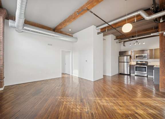 A room with wooden floors and white walls, with a kitchen area visible in the background.